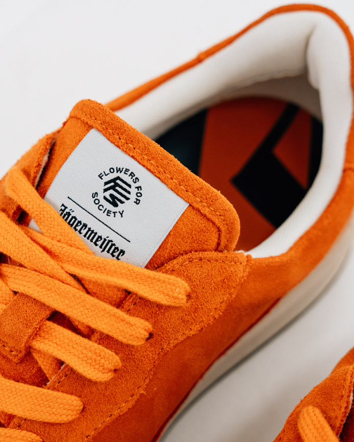 Close-up of an orange Jägermeister sneaker with matching orange laces, a white tongue label reading 'Flowers For Society' and 'Jägermeister', and a white side stripe on a white background.