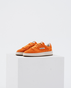 A pair of orange Jägermeister sneakers with white midsoles and gum soles, displayed on a white geometric pedestal against a light gray background.