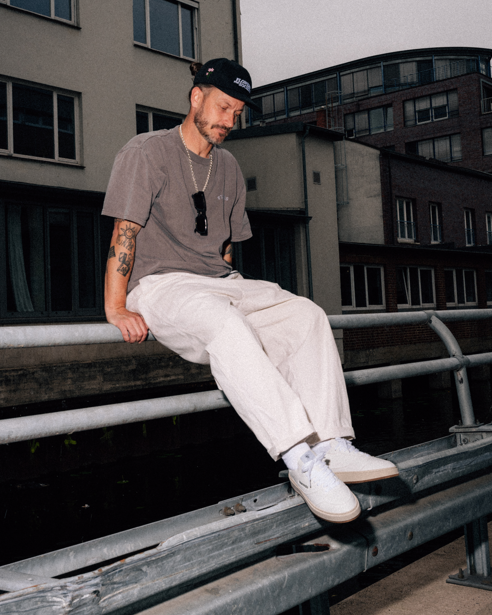A man wearing a black baseball cap, taupe t-shirt, off-white trousers, and white Jägermeister sneakers sits on a metal guardrail in an urban outdoor setting.