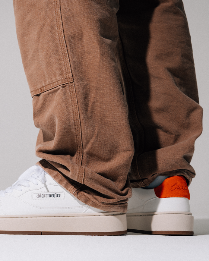 Close-up of a person wearing brown corduroy pants and white Jägermeister sneakers, with the back heel of one shoe showing a bright orange heel tab and black script Jägermeister branding.