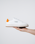 A white Jägermeister sneaker with a bright orange heel tab and a black 'Jägermeister' wordmark on the side, held up by a person's hand against a light background.
