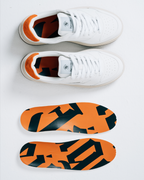 A pair of white Jägermeister sneakers shown from the top, with two matching insoles displayed below. The insoles feature a bold orange and black abstract Jägermeister wordmark graphic pattern.
