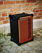 Haut-parleur portable Soundboks orange et noir avec la marque « Jägermeister » devant un mur de briques