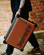 Une personne tient un haut-parleur portable Soundboks orange et noir avec la marque « Jägermeister » devant un mur de briques.