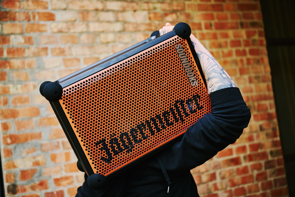 Une personne tient un haut-parleur portable Soundboks orange et noir avec la marque « Jägermeister » devant un mur de briques.
