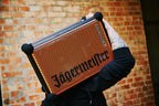 Une personne tient un haut-parleur portable Soundboks orange et noir avec la marque « Jägermeister » devant un mur de briques.