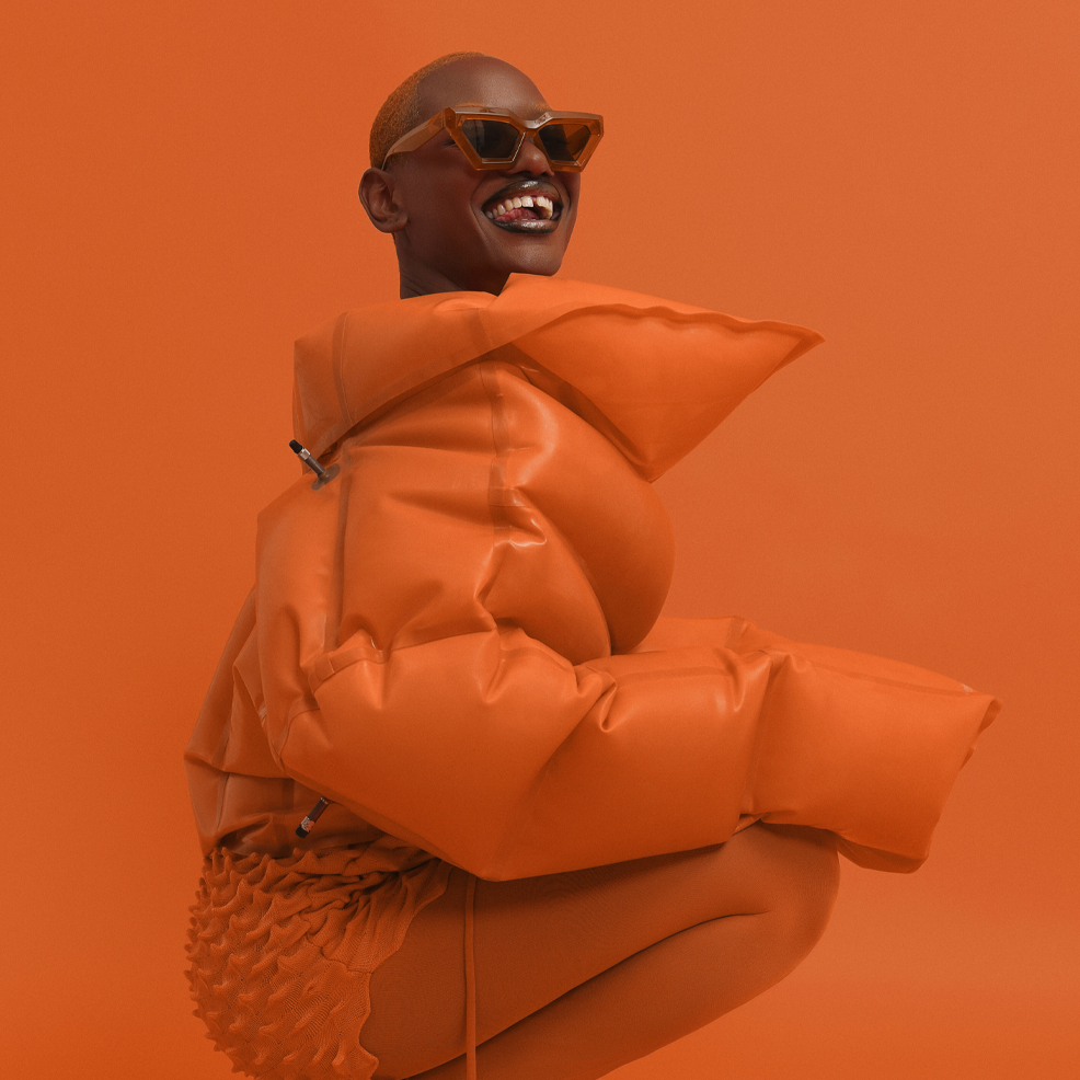 A model wearing a chunky, oversized orange puffer jacket with a high structured collar and matching textured orange bottoms, posing against a solid orange studio background.