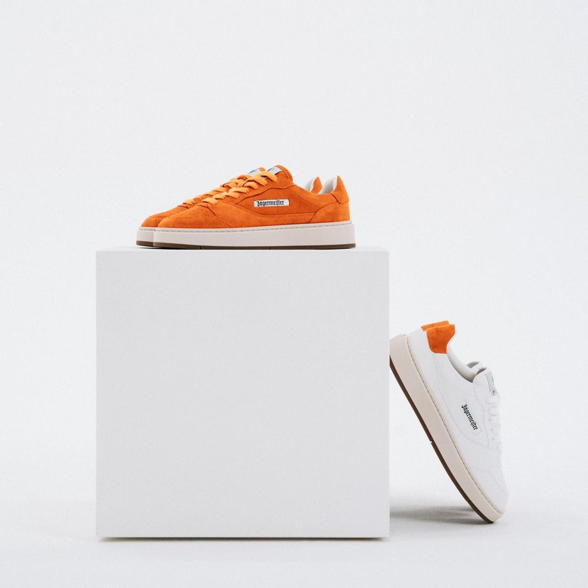 A pair of sneakers displayed on a white pedestal against a light gray studio background. One shoe is bright orange suede with a white sole, and the other is white suede with an orange heel tab and white sole.