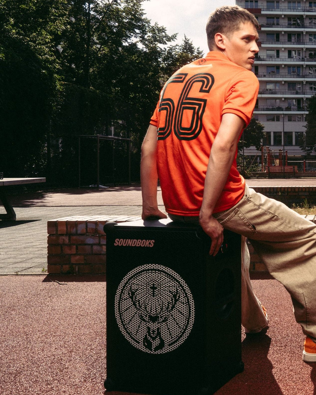 Un homme en maillot Jägermeister orange et pantalon beige est assis sur un haut-parleur Soundboks noir avec le logo de la marque Jägermeister
