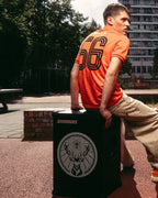 Un homme en maillot Jägermeister orange et pantalon beige est assis sur un haut-parleur Soundboks noir avec le logo de la marque Jägermeister