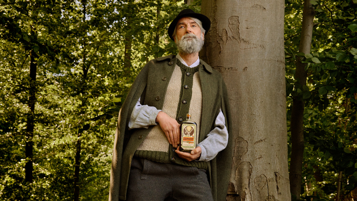 A man with a gray beard and black hat stands outdoors against a large tree trunk in a wooded setting, wearing a dark green cape over a beige knit sweater and light blue shirt, while holding a retro-bottle of Jägermeister.