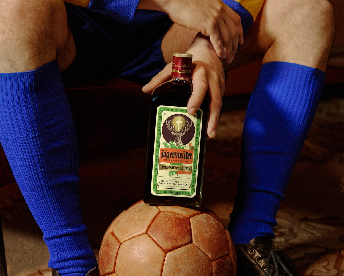 A person in a yellow and blue soccer jersey sits on a bench with legs crossed, holding a Jägermeister retro-bottle. A vintage soccer ball rests on their shins, and they are wearing blue knee-high socks and black leather soccer boots.