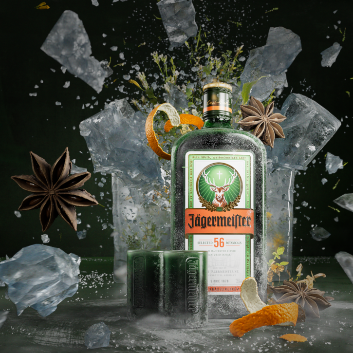 A bottle of Jägermeister with two small tumblers, surrounded by dramatic ice cubes, star anise, orange peel, and botanicals against a dark background.