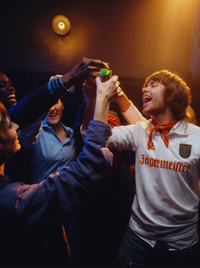 Groupe de personnes célébrant avec une personne portant un t-shirt Jägermeister dans une pièce faiblement éclairée.